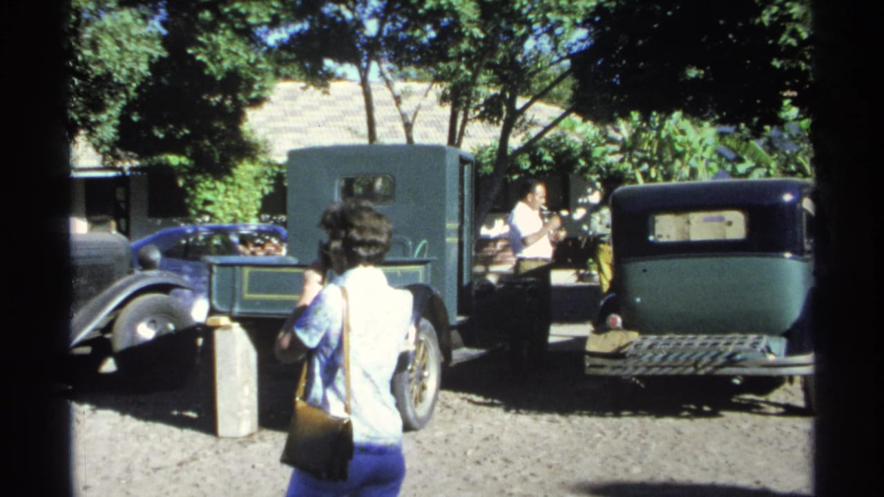 Older green car and dark sedan parked in a courtyard as adults stand nearby.