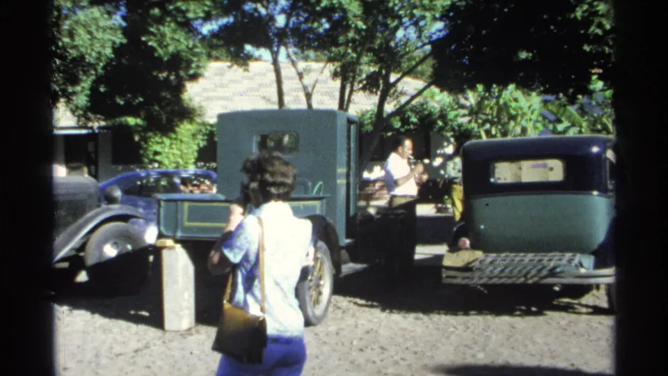 A 1970s home-movie frame shows adults near older parked cars in a bright Guaymas courtyard shaded by trees.