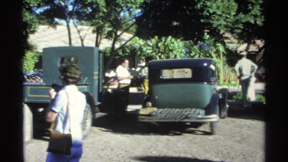 The contact frame shows a woman at left and a dark older sedan centered in a gravel courtyard, with adults conversing in the background.