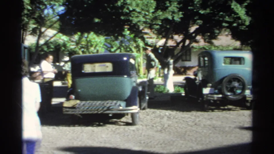 A dark sedan and a light blue older car are parked in a Guaymas courtyard as adults stand and talk under trees.