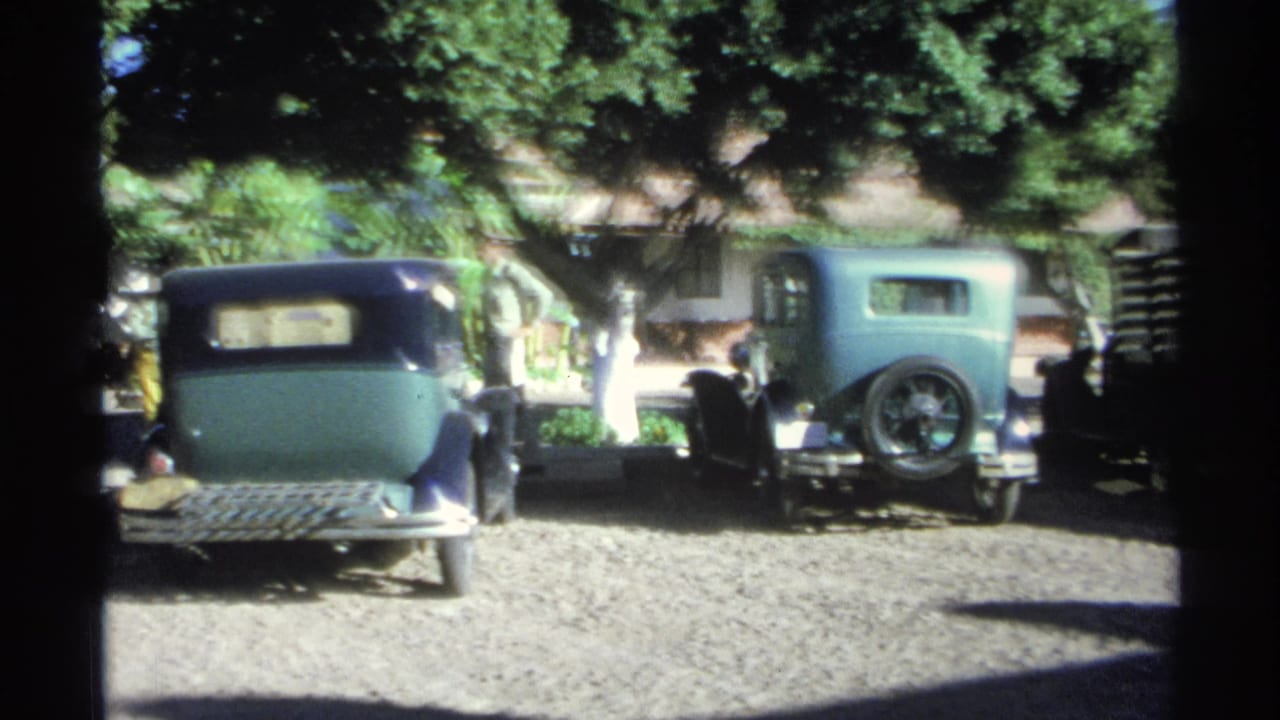 1975 Guaymas home-movie frame with older cars parked under leafy trees on a sunlit gravel street area.