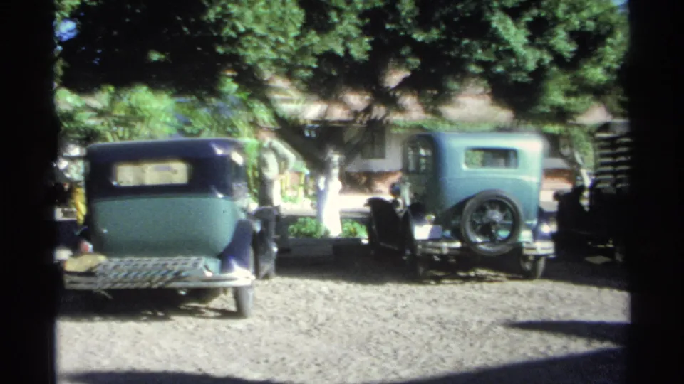 1975 Guaymas home-movie frame with older cars parked under leafy trees on a sunlit gravel street area.