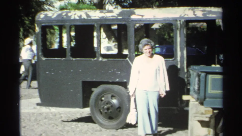 1975 Guaymas home-movie frame with a woman walking in front of a dark bus on a sunlit gravel street.