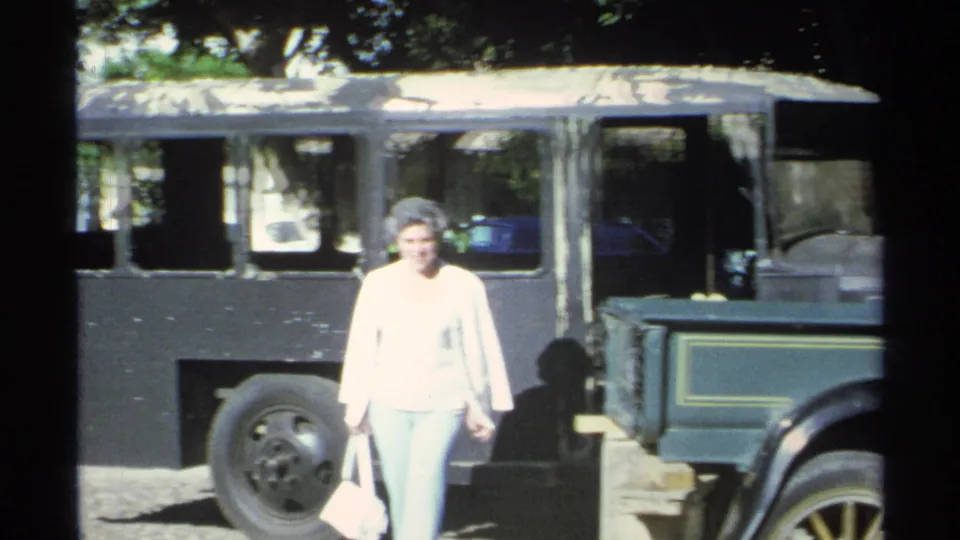 1975 Guaymas frame with a woman walking near a dark bus, street fixture, and partial older vehicle.
