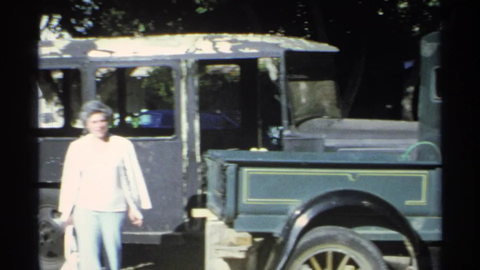 In Guaymas, Mexico, 1975, a woman stands beside a green vintage pickup with yellow trim in shaded 8mm footage.