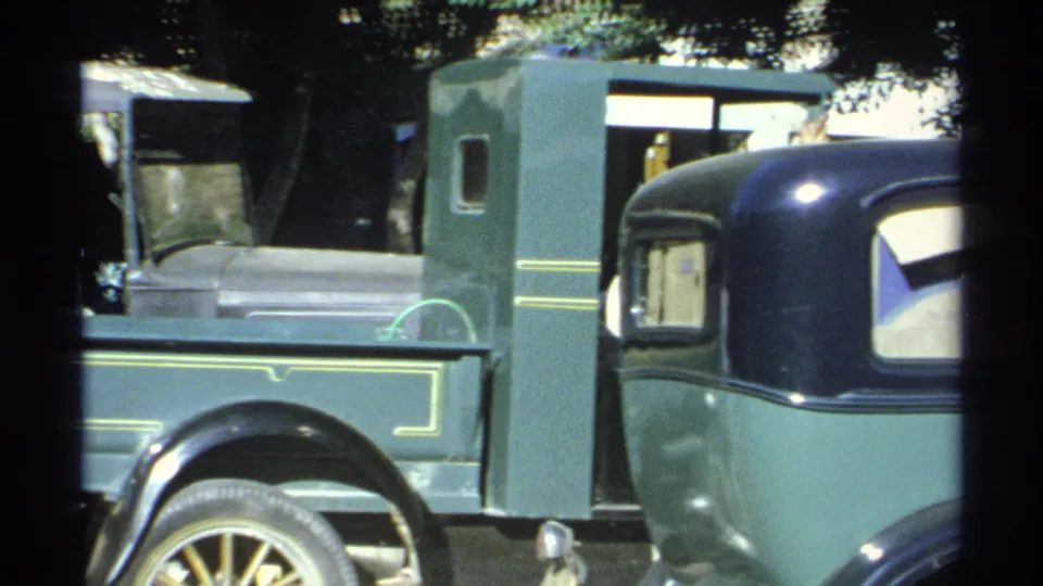 A green vintage pickup with spoked wheels and yellow trim is seen closely, with another car entering the frame at right.