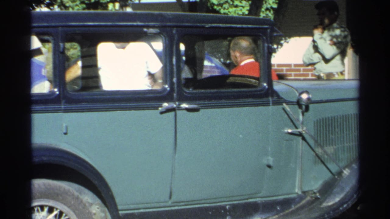 Green and black vintage sedan with people visible inside, parked outdoors in sun.