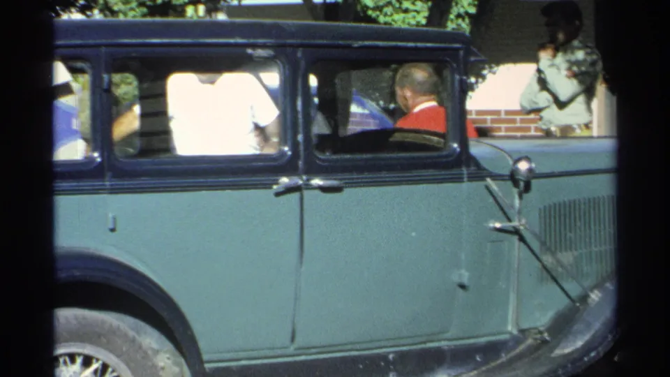 1975 Guaymas home-movie frame showing a green and black vintage sedan with occupants, trees, and a brick wall behind it.