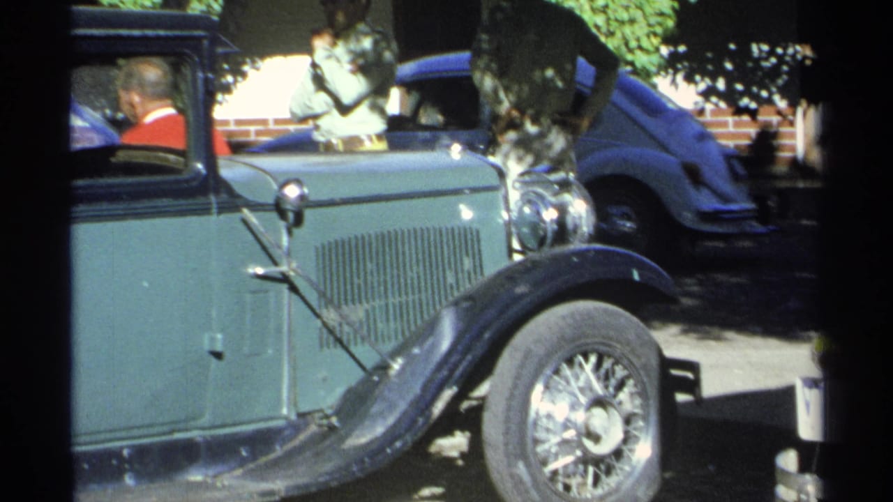 Side view of the green and black vintage automobile in Guaymas, Mexico, with a seated driver and nearby parked vehicle partly visible.