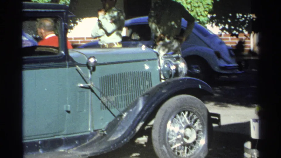 Side view of the green and black vintage automobile in Guaymas, Mexico, with a seated driver and nearby parked vehicle partly visible.