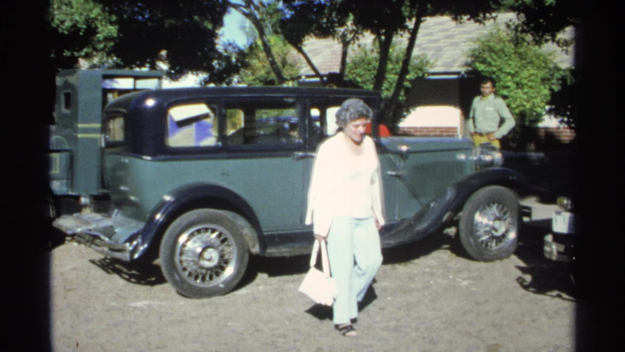 A woman in a light top and pants walks past the green vintage sedan during a 1975 Guaymas home-movie street scene.