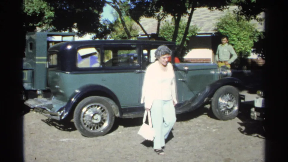 A woman in a light top and pants walks past the green vintage sedan during a 1975 Guaymas home-movie street scene.