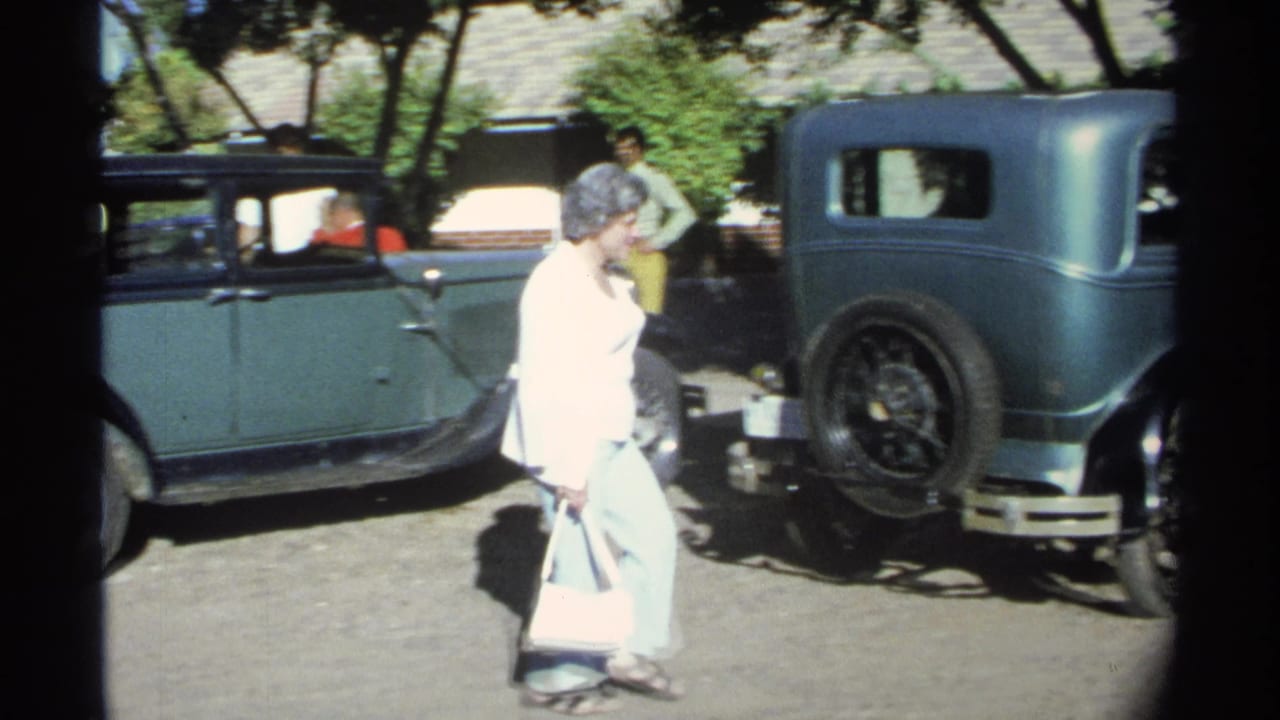 The woman continues walking past the green sedan, with another dark-fendered vintage car and the rear of the green car visible nearby.