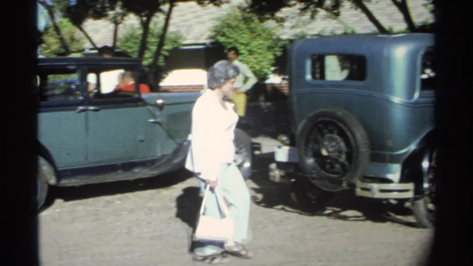 The woman continues walking past the green sedan, with another dark-fendered vintage car and the rear of the green car visible nearby.