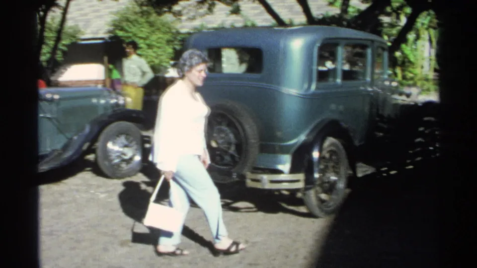 In Guaymas, Mexico in 1975, a woman with a light handbag walks past a teal vintage sedan and another parked car in bright sun.