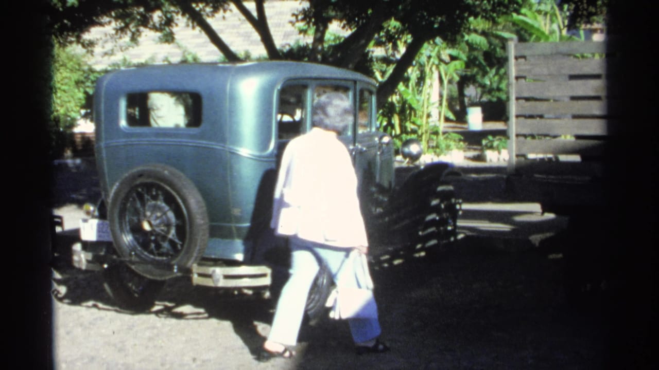 The woman walks toward the passenger side of a teal older sedan in Guaymas, with trees, a fence, and strong daylight around the parked car.