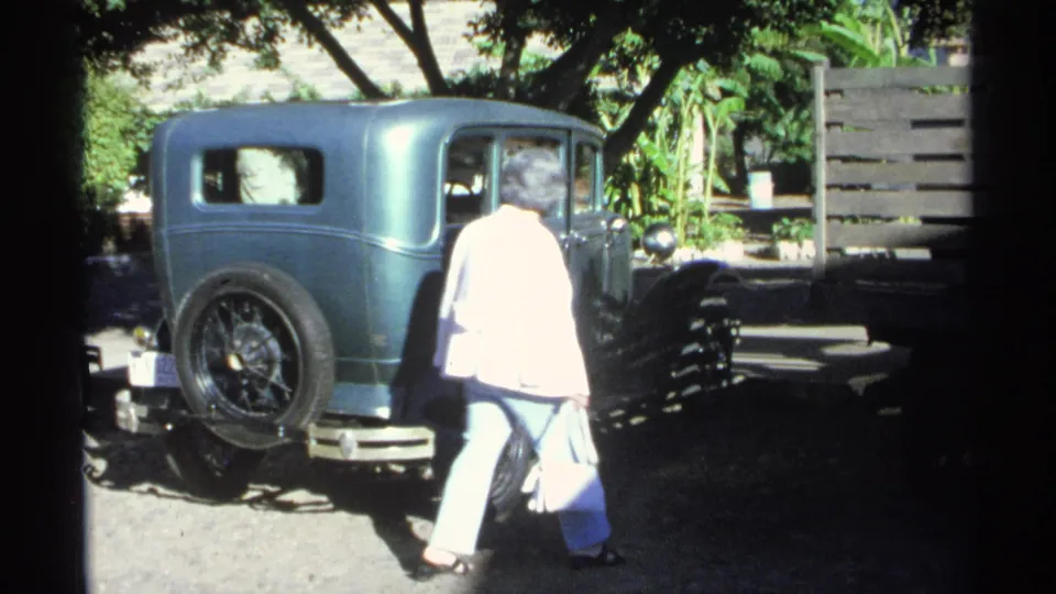 The woman walks toward the passenger side of a teal older sedan in Guaymas, with trees, a fence, and strong daylight around the parked car.