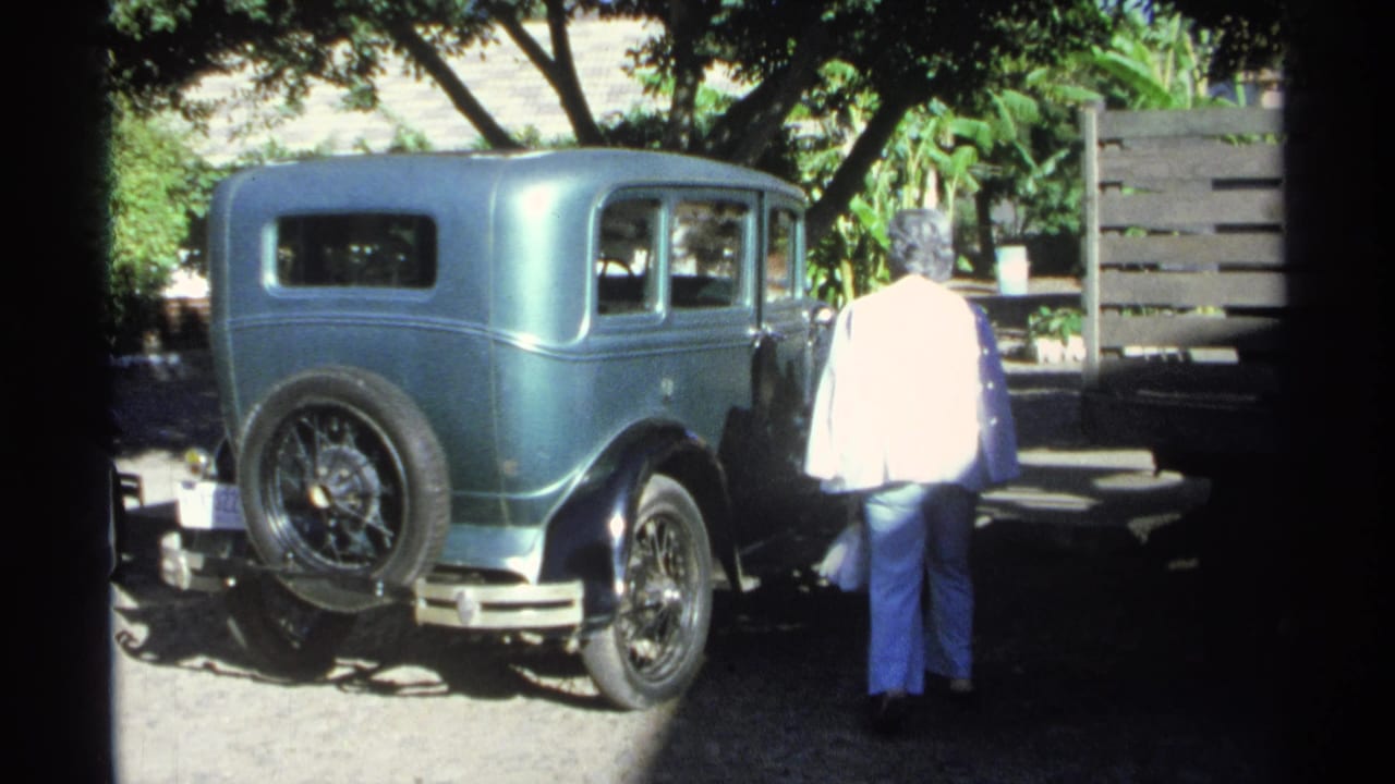 A woman pauses beside a teal vintage sedan in the 1975 Guaymas travel footage, standing near the side windows in a shaded parking area.