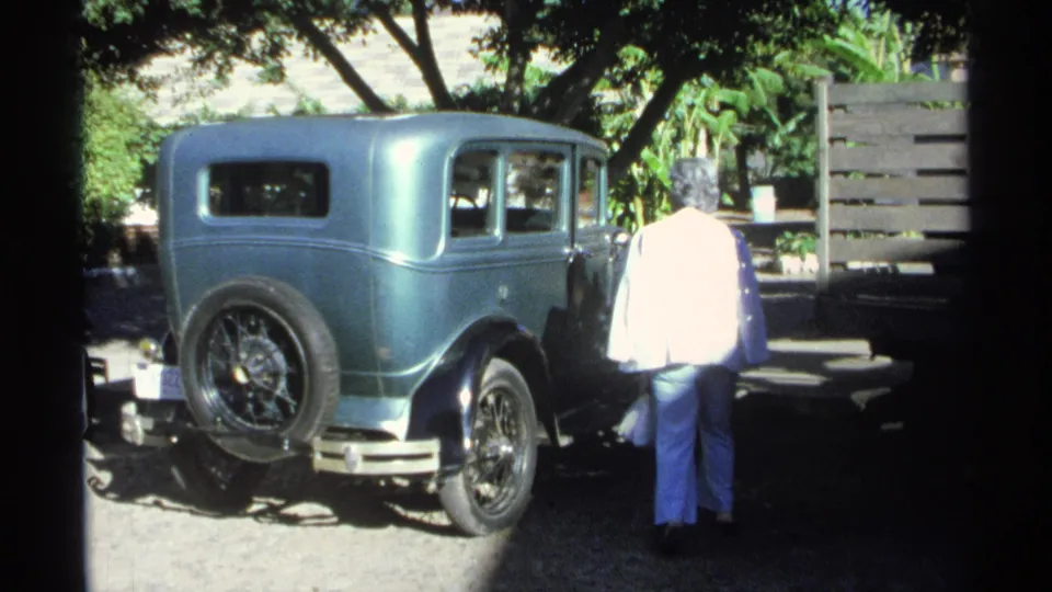 A woman pauses beside a teal vintage sedan in the 1975 Guaymas travel footage, standing near the side windows in a shaded parking area.