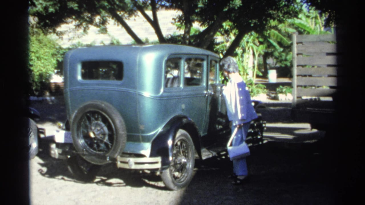 The woman appears to look into the side window of a teal vintage sedan during a 1975 Guaymas vacation scene, with strong sun and shade across the frame.