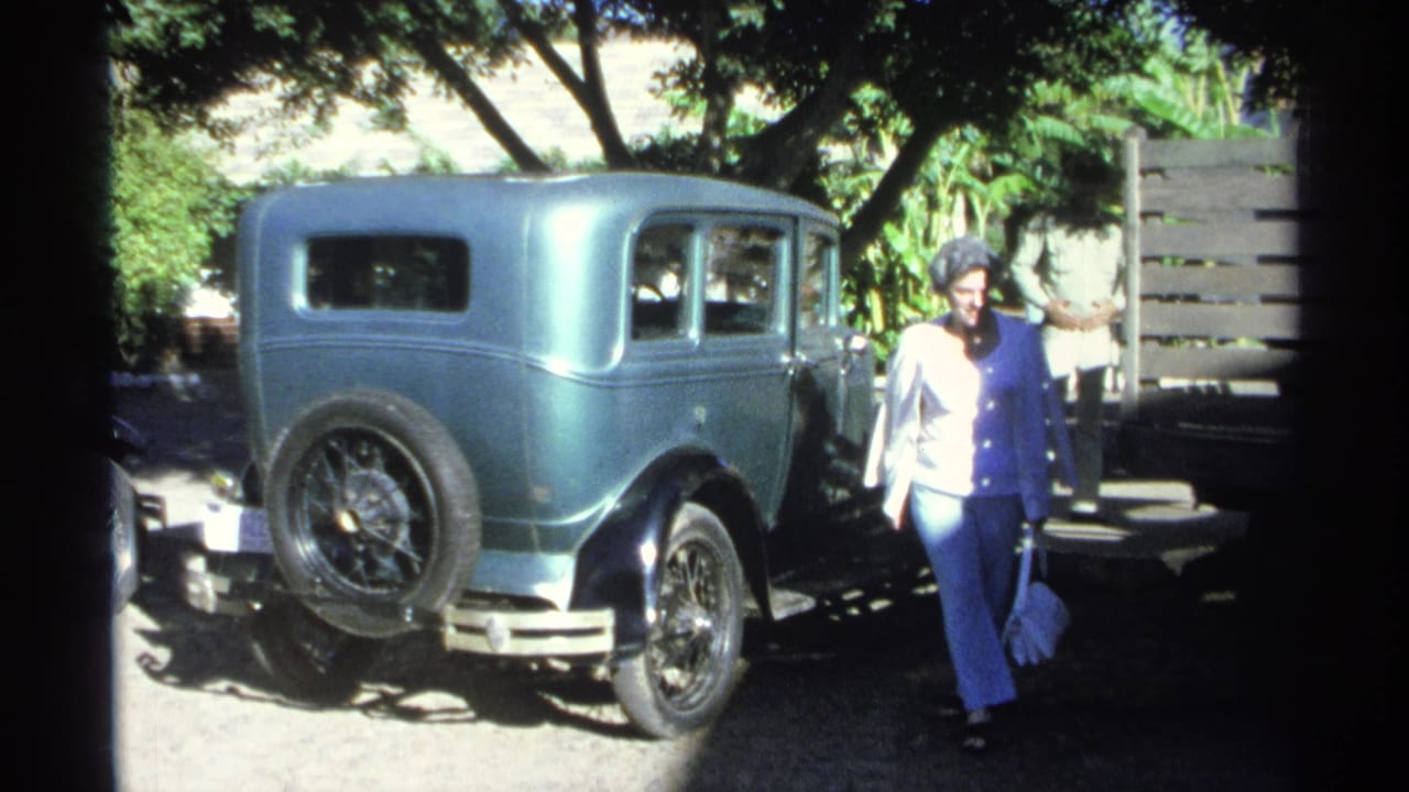 A woman walks near the rear of the turquoise vintage automobile, with trees and a wooden fence behind her in Guaymas.