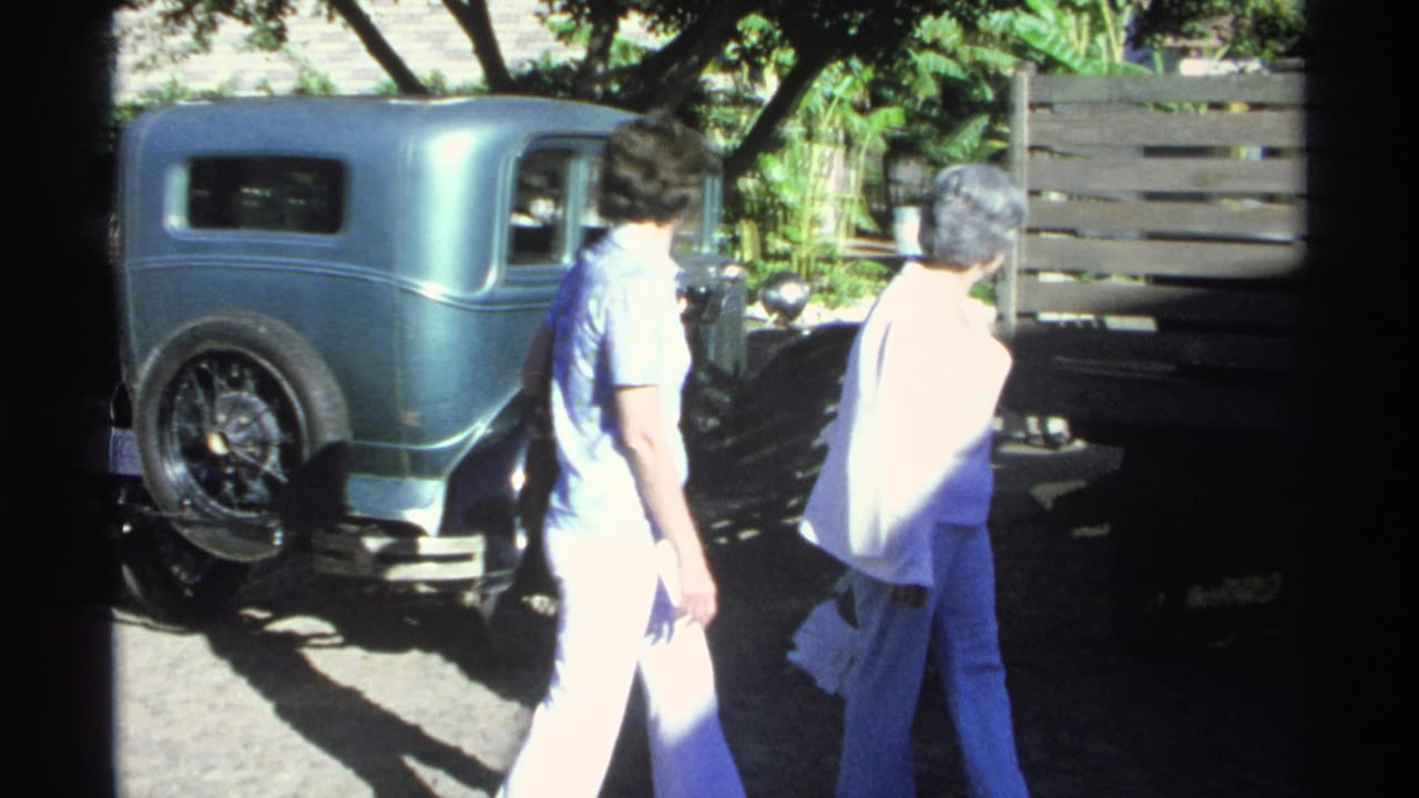 Two adults appear beside the turquoise vintage car in the 1975 Guaymas home-movie scene, with strong sunlight and tree shade.