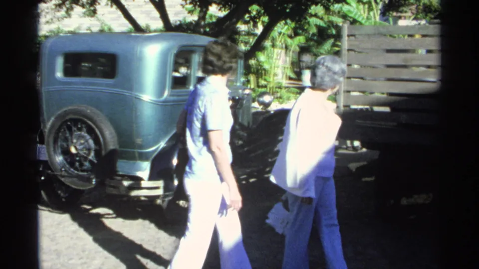 Two adults appear beside the turquoise vintage car in the 1975 Guaymas home-movie scene, with strong sunlight and tree shade.