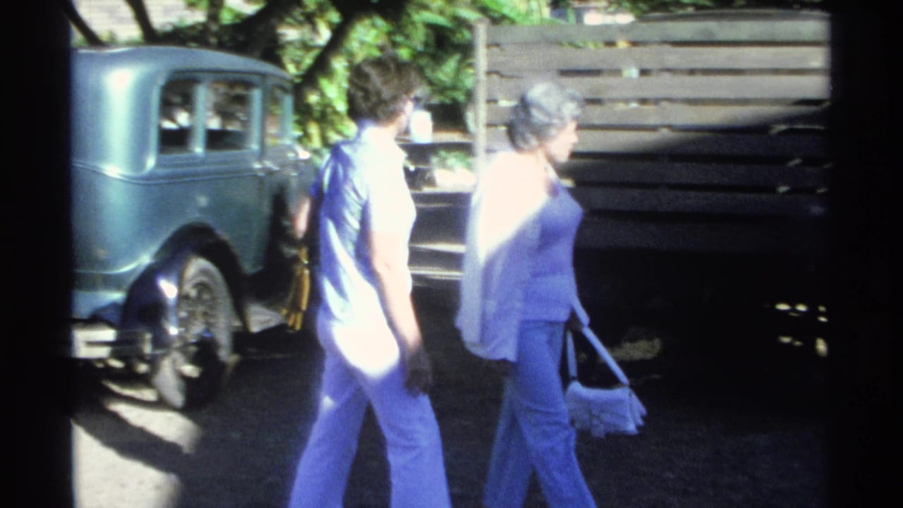 In Guaymas, Mexico, 1975, two women in light clothing walk beside an older blue sedan near trees and a wooden fence.