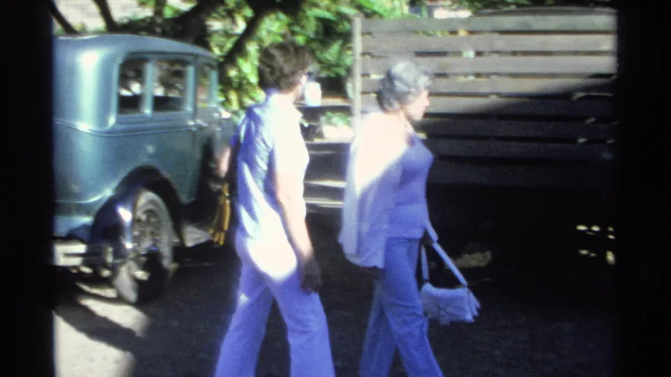 In Guaymas, Mexico, 1975, two women in light clothing walk beside an older blue sedan near trees and a wooden fence.
