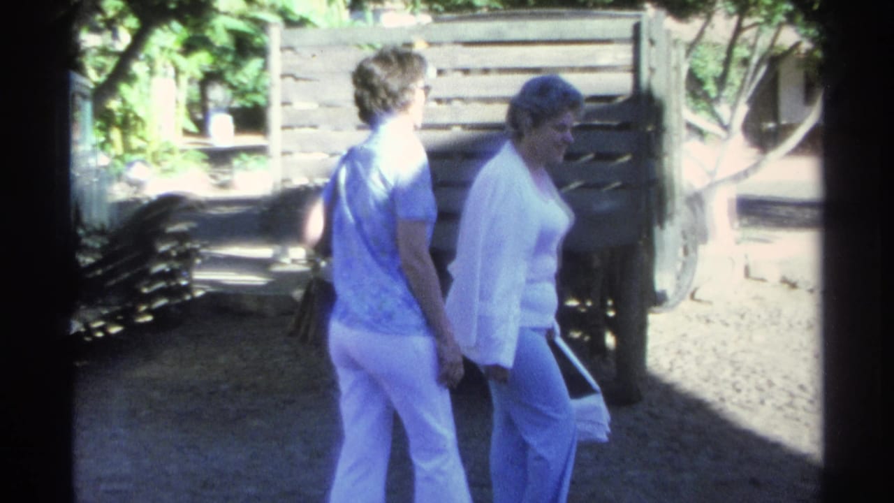 Two women walk past a wooden-sided truck in bright Guaymas sunlight, with foliage and fencing behind them.
