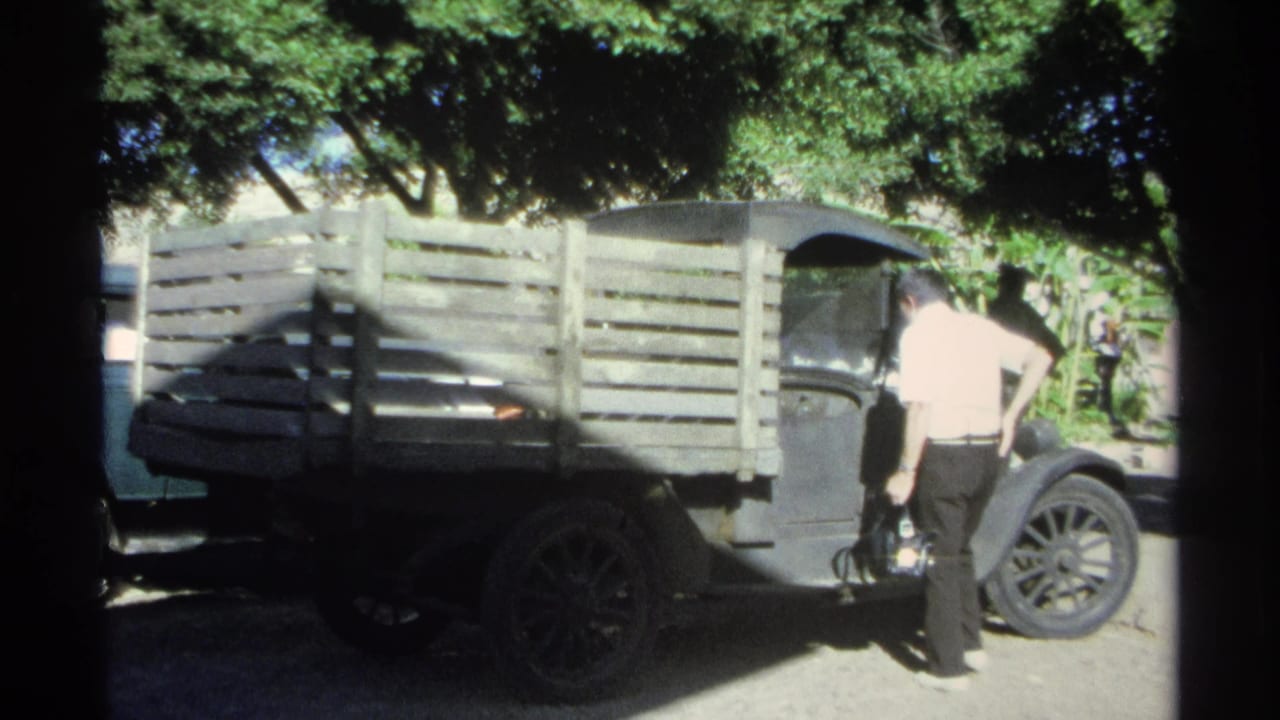 A man in a light shirt stands beside the cab of a wooden-sided truck, with trees and a fence in the background.