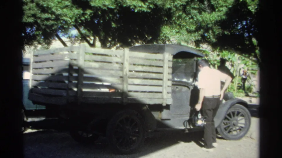 A man in a light shirt stands beside the cab of a wooden-sided truck, with trees and a fence in the background.