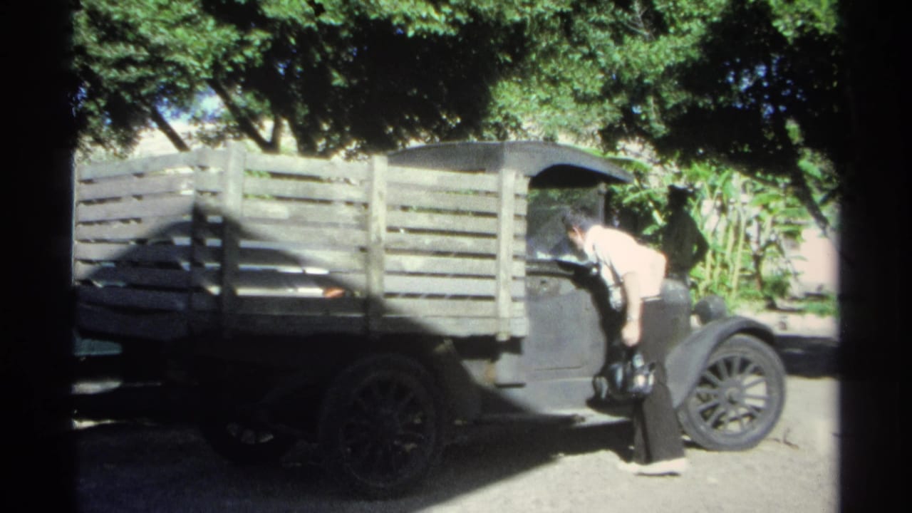 The man leans near the front cab area of the wooden-sided truck in a shaded, tree-lined outdoor space.