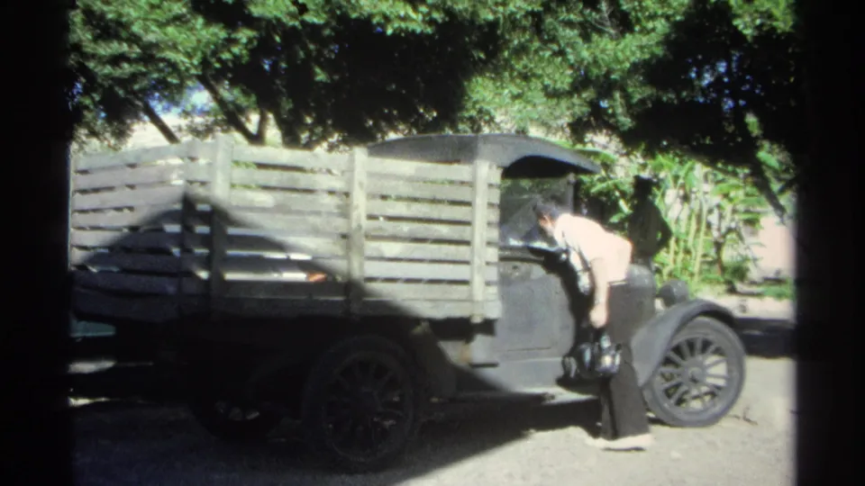 The man leans near the front cab area of the wooden-sided truck in a shaded, tree-lined outdoor space.