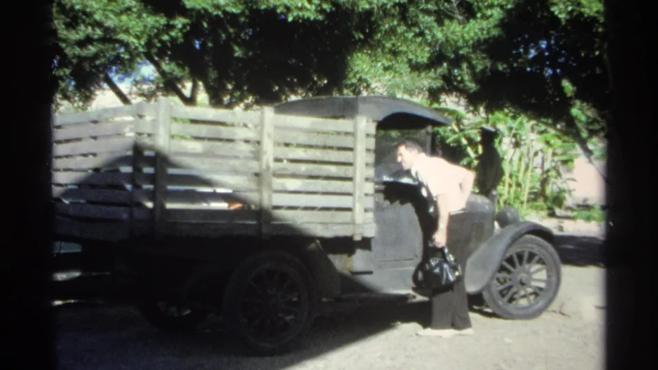 In 1975 Guaymas, Mexico, a man in a white shirt bends near the driver's side of a dark pickup truck with wooden side slats.