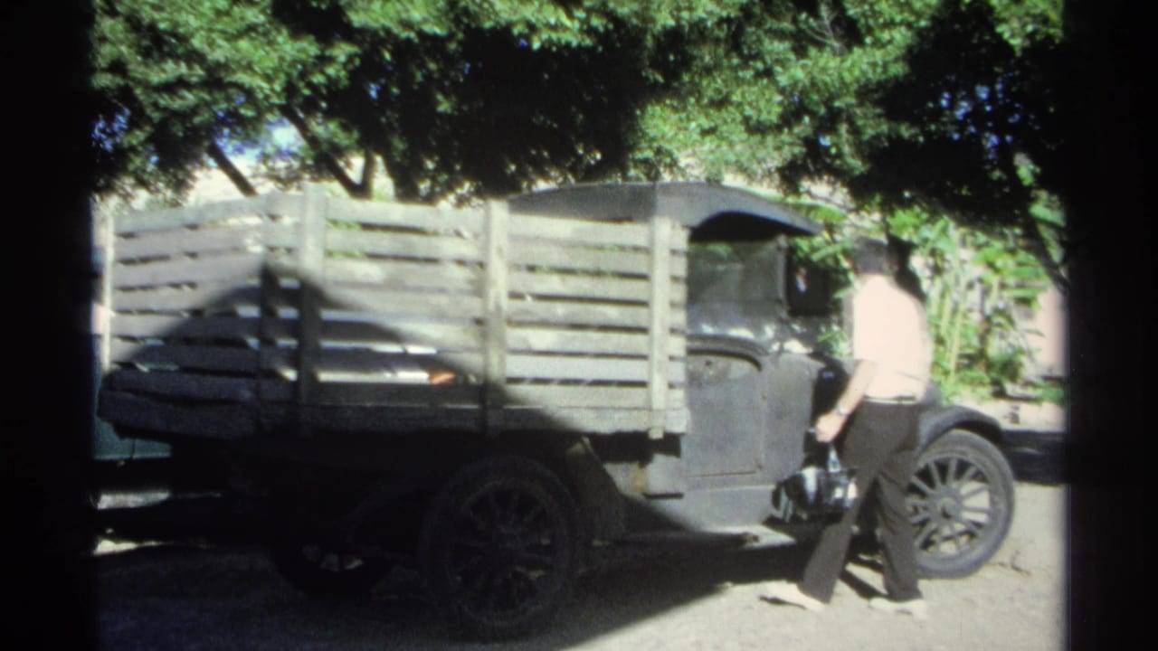 A man in a light shirt stands near the driver's door of a dark wooden-bed pickup in Guaymas, Mexico, during the 1975 home movie footage.