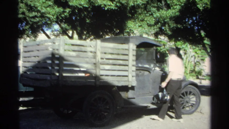 A man in a light shirt stands near the driver's door of a dark wooden-bed pickup in Guaymas, Mexico, during the 1975 home movie footage.