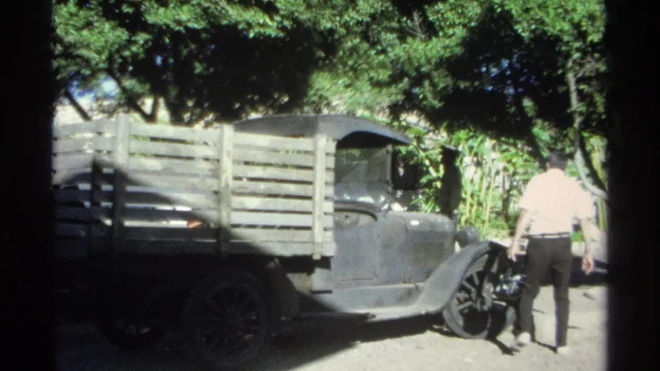 The man is seen from behind moving along the driver's side of the parked black pickup, with leafy shade and sunlit dirt around the vehicle.