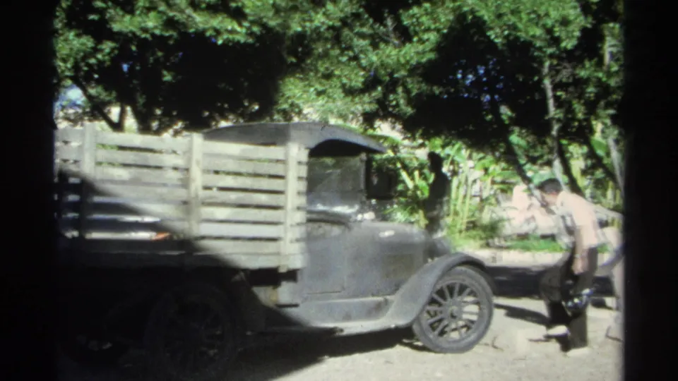 In 1975 Guaymas footage, adult men examine a wooden-slat flatbed truck parked in shade beneath leafy trees.