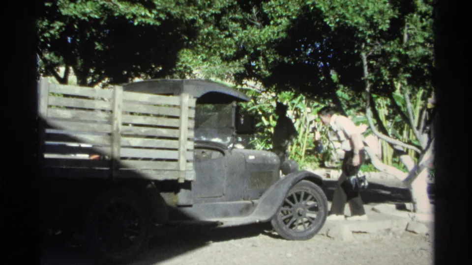 An adult man bends near the front fender of a wooden-slat flatbed truck in 1975 Guaymas home-movie footage.