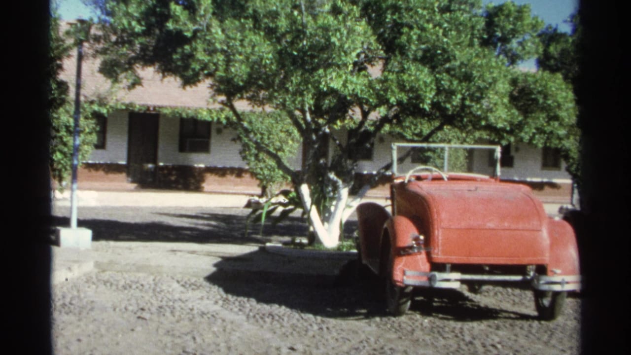 A red vintage roadster sits on gravel near leafy trees and a stucco building in 1975 Guaymas travel footage.