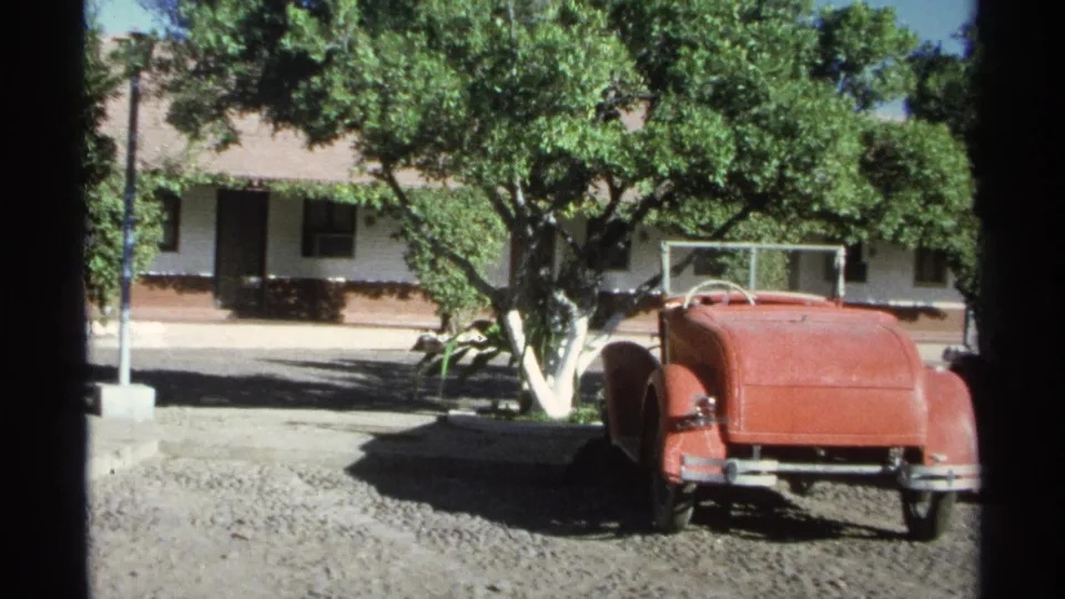 A red vintage roadster sits on gravel near leafy trees and a stucco building in 1975 Guaymas travel footage.