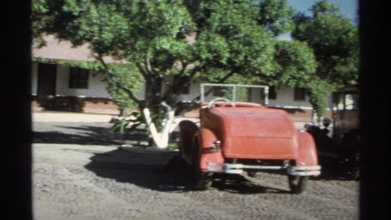 1975 Guaymas resort parking view with a red vintage convertible on gravel beside trees and a low stucco building.