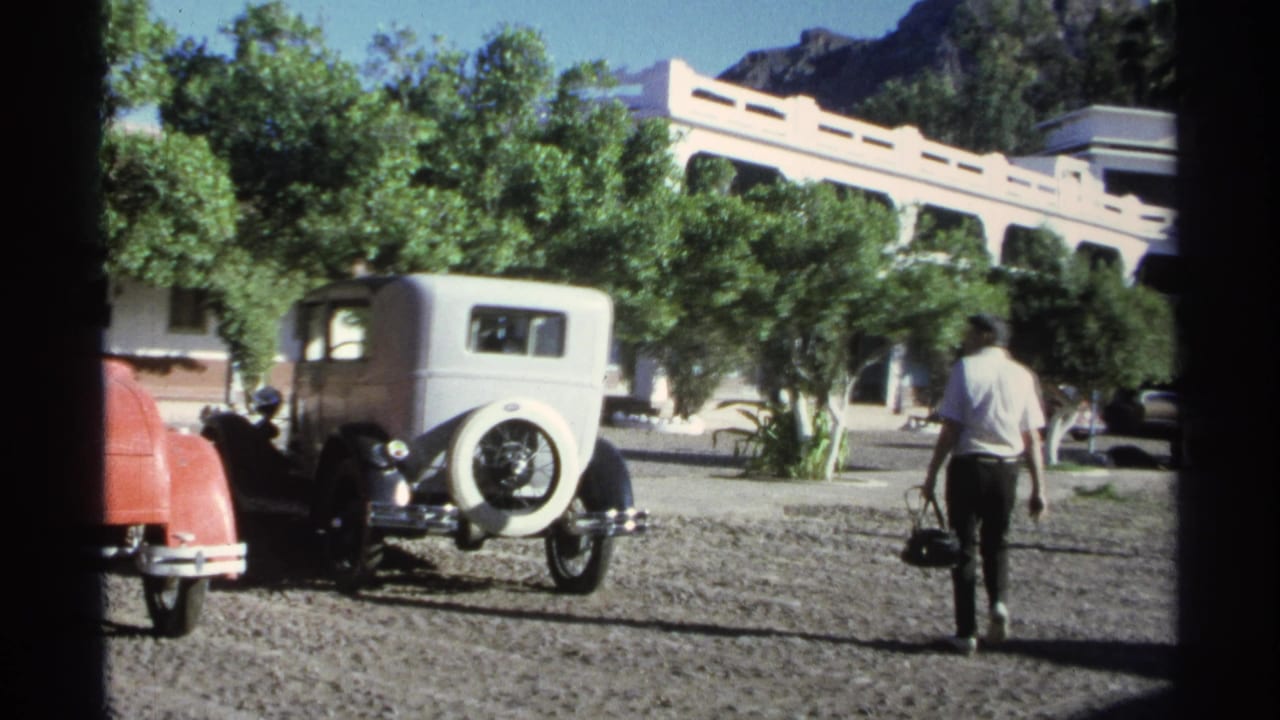 1975 Guaymas resort scene with a man carrying a bag past parked vintage cars, trees, and a low building.