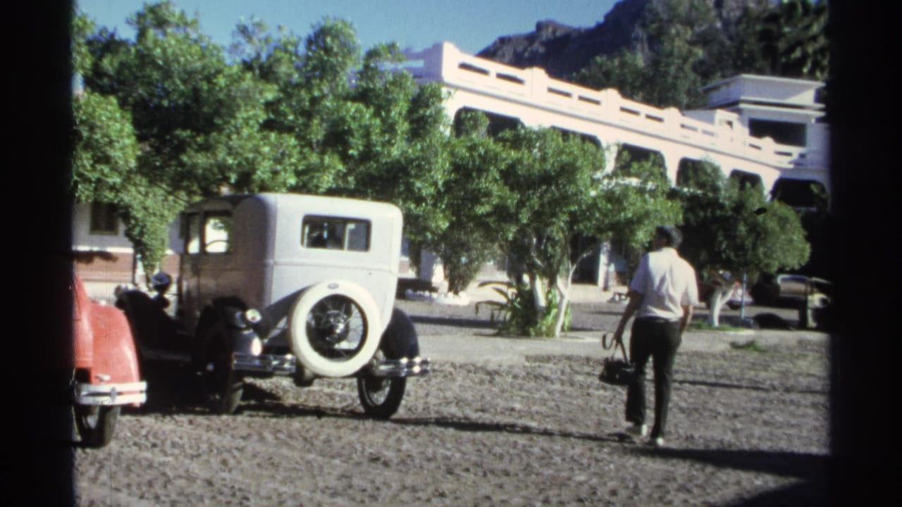 In Guaymas, Mexico in 1975, a man carrying a black bag walks across gravel near a white vintage car and a pale arched building.