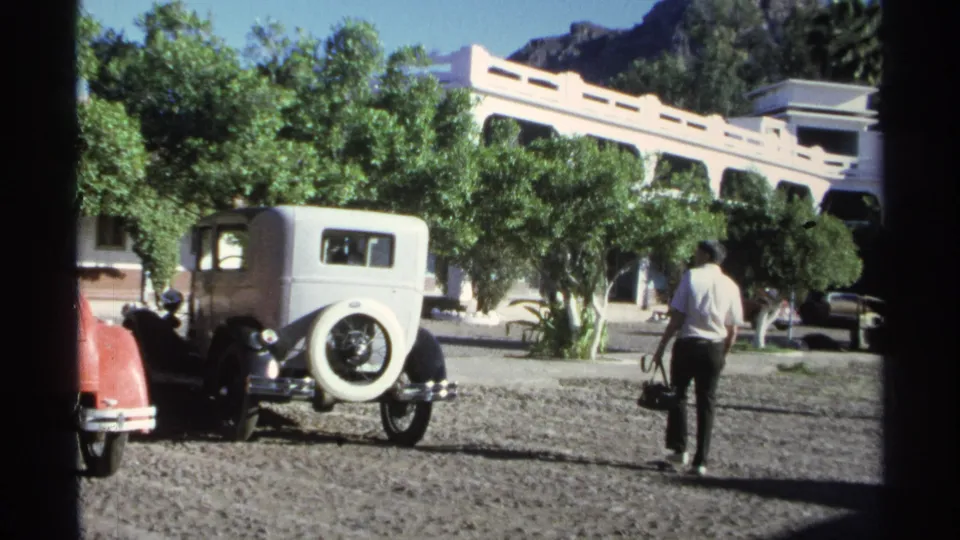 In Guaymas, Mexico in 1975, a man carrying a black bag walks across gravel near a white vintage car and a pale arched building.