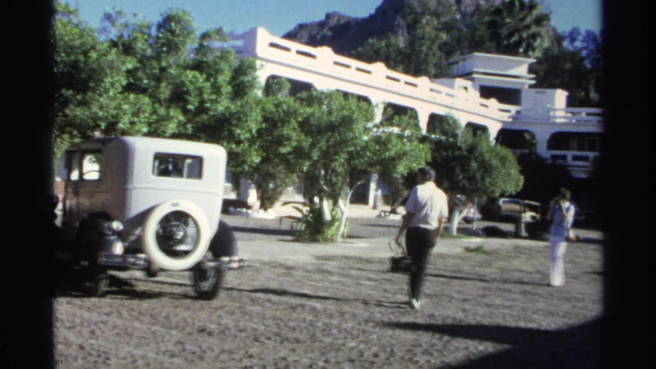 Man walks away from the camera near a white vintage car and leafy trees.