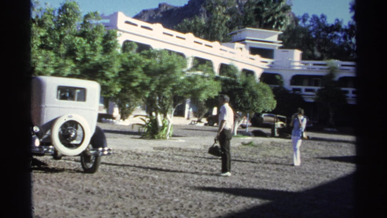 The man carrying a black bag passes through a sunlit Guaymas courtyard with a white vintage car, trees, and another person in the distance.