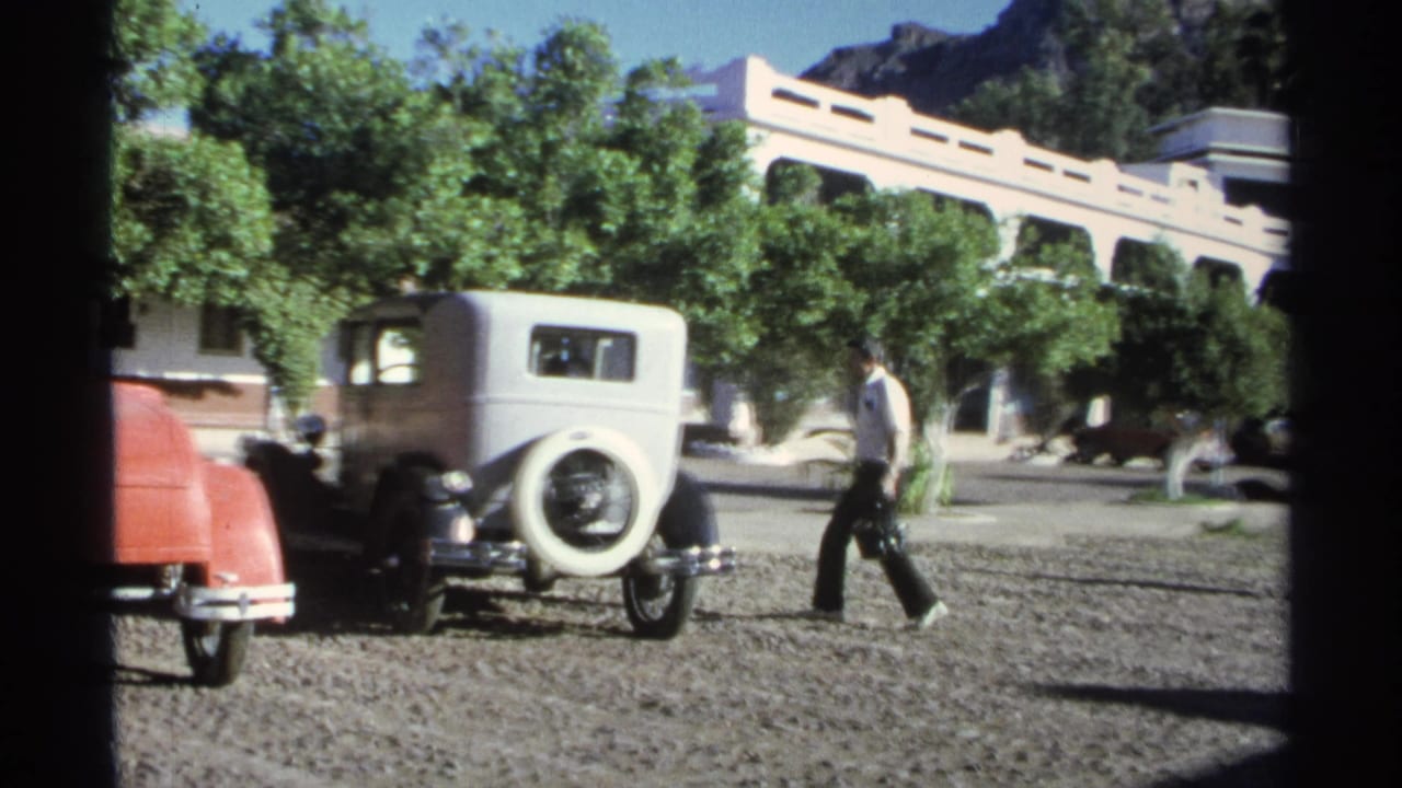 Man walking near white vintage sedan and red car in a gravel courtyard.