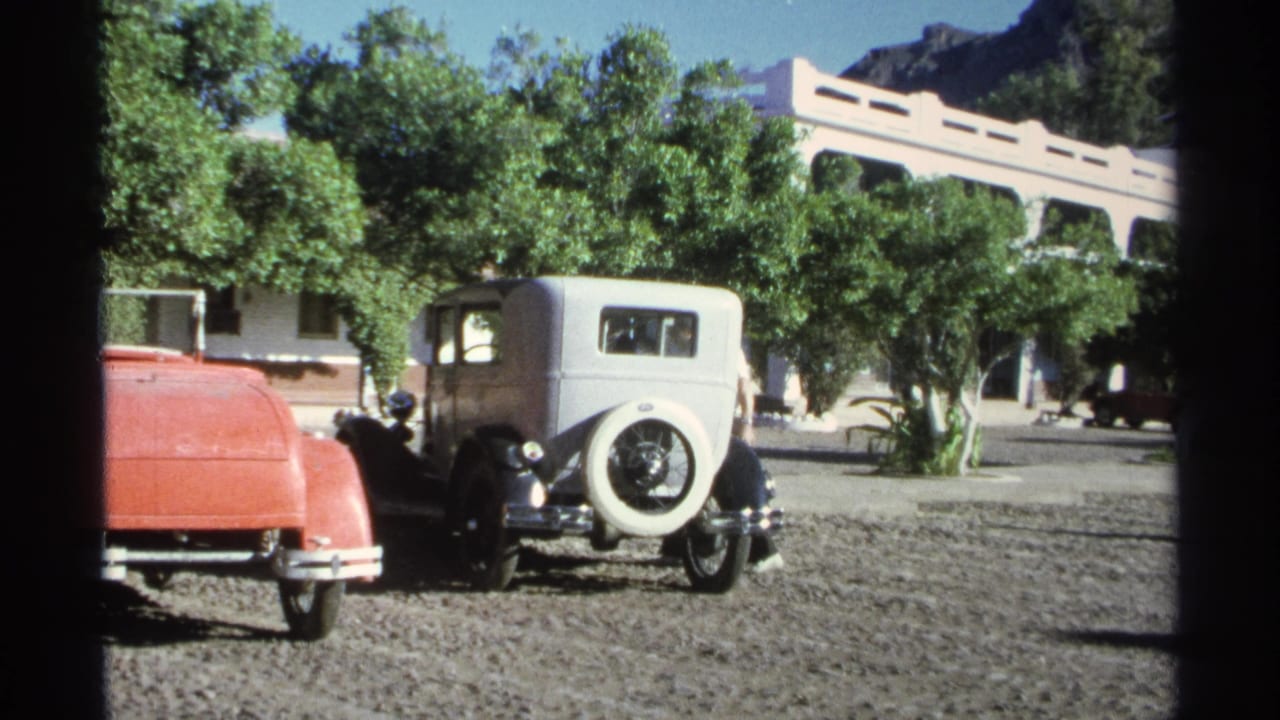 1975 Guaymas frame showing a white sedan with rear spare tire parked near a red car, trees, and a white arched building.